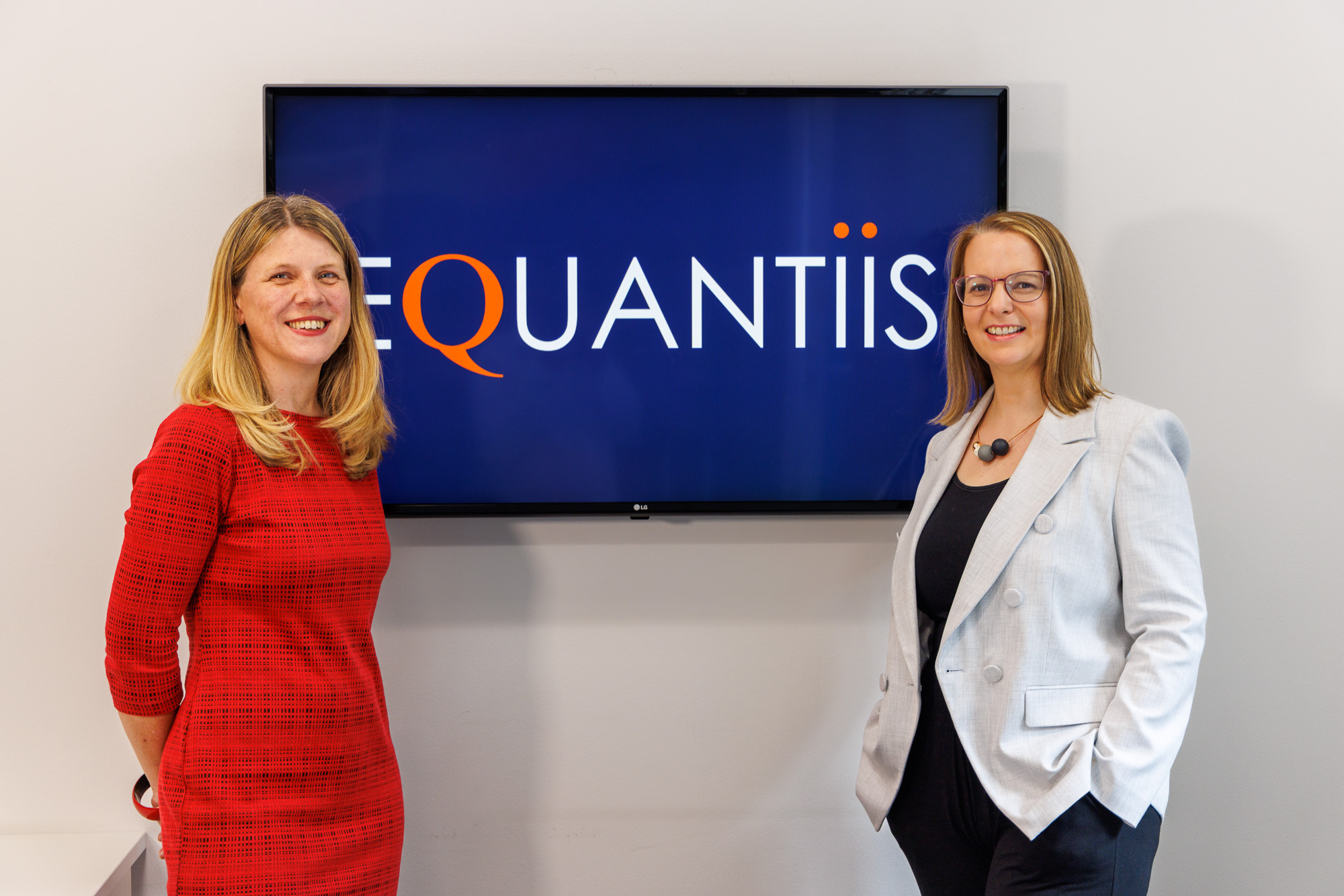 two female equantiis consultants standing next to a tv with company logo on screen