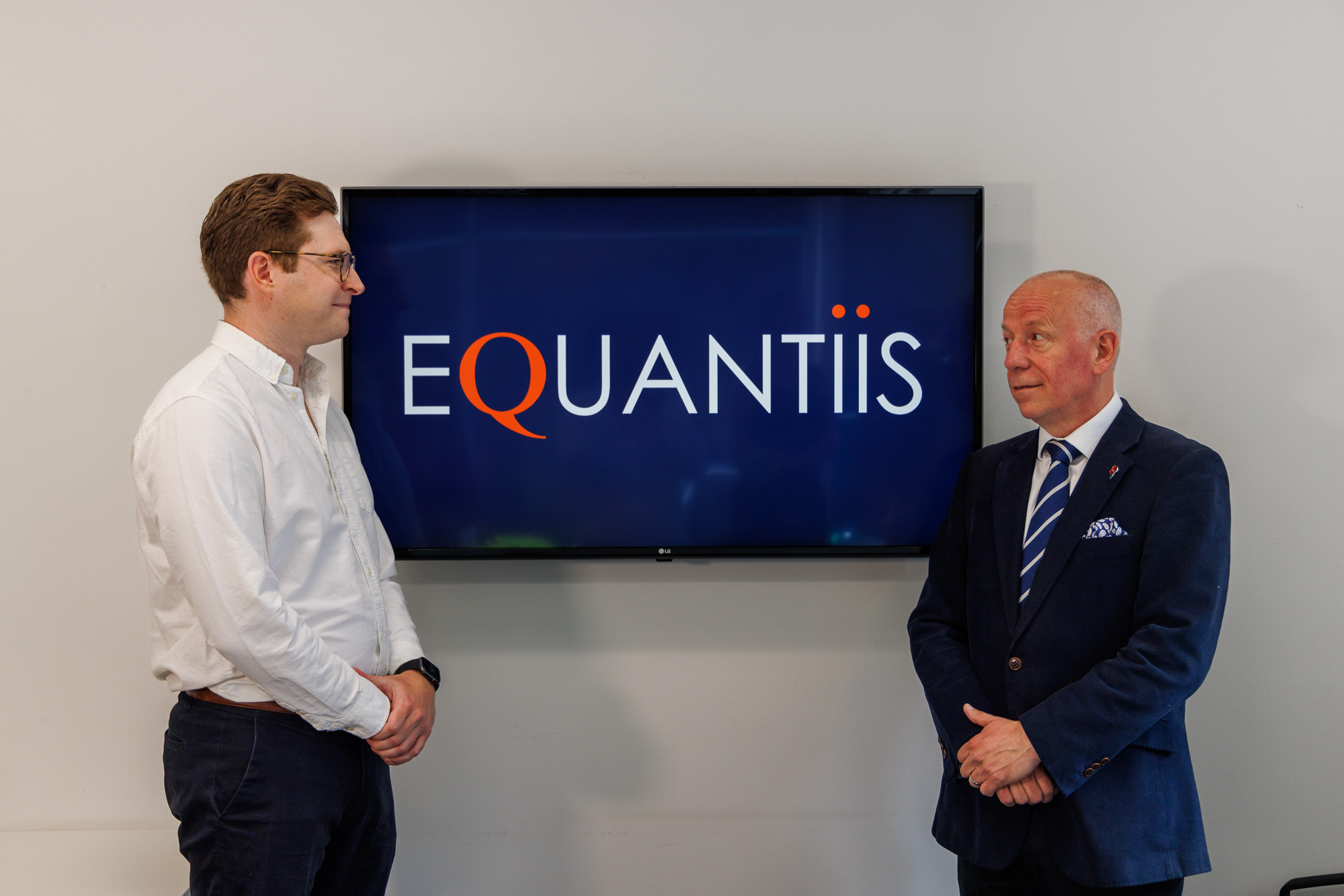 two equantiis consultants standing next to a tv with company logo on screen