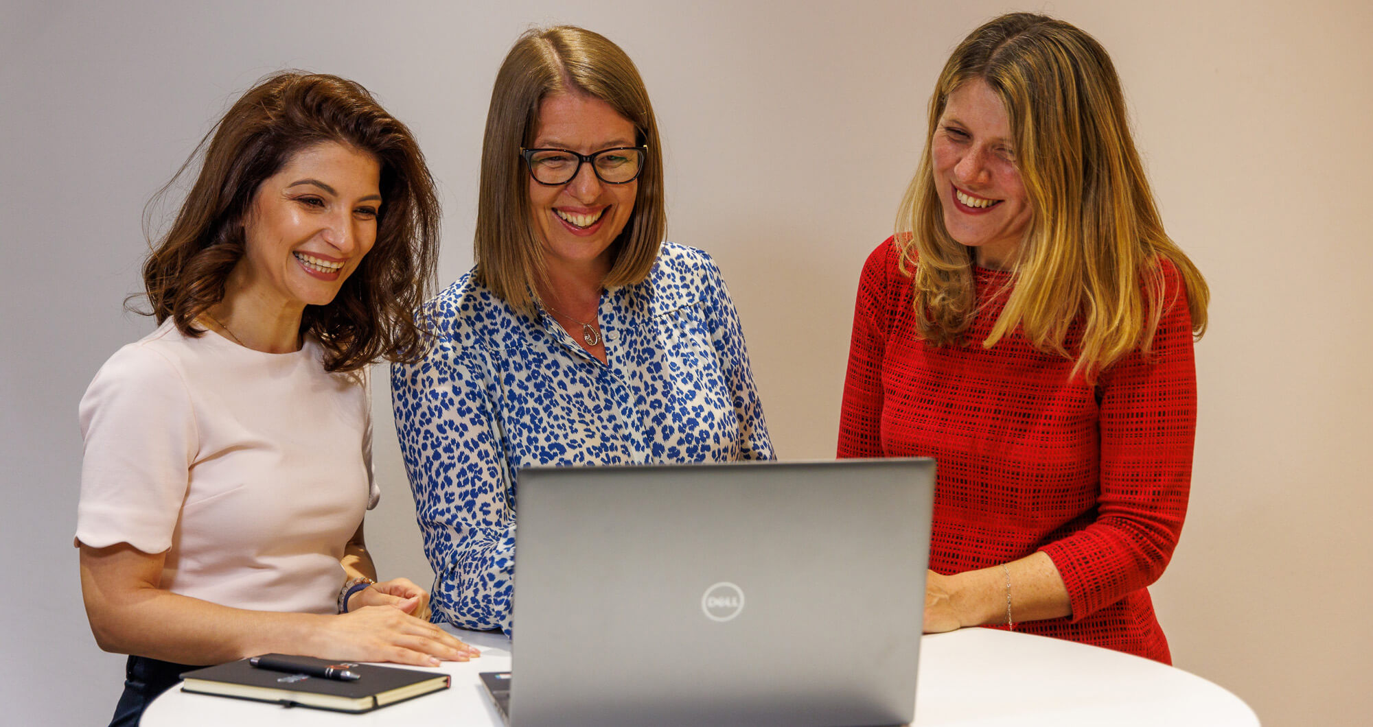 3 women standing around a laptop, watching the screen,smiling